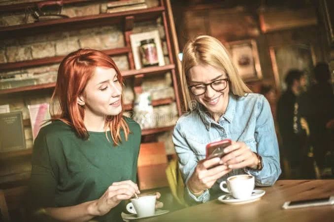 Funny Shayari On Girls Attitude showing two women sharing a smartphone and laughing at a cafe