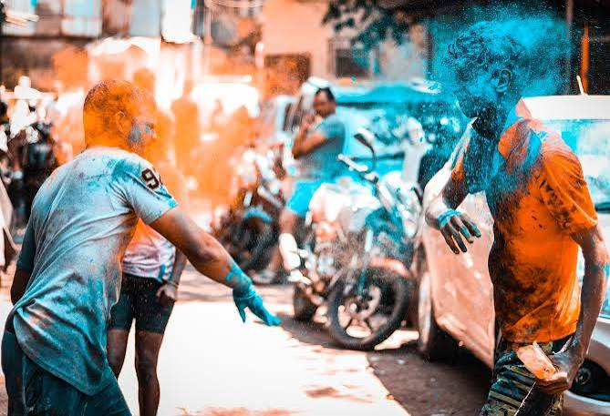 Holi Sad Shayari In Hindi showing people celebrating Holi with colorful powders and a festive street atmosphere